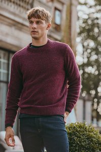 Jonge man in een bordeauxrode trui en donkere spijkerbroek, staand in de buitenlucht nabij groene struiken en een gebouw.