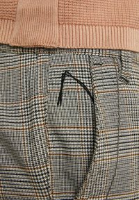 Béžový pletený svetr zastrčený do šedých a hnědých kostičkovaných kalhot s černou výšivkou poblíž kapsy.