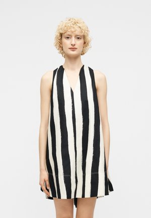 Jeune personne aux cheveux blonds bouclés et courts, portant une robe sans manches à rayures verticales noires et blanches avec un col en V, debout devant un fond uni.