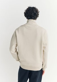 Beige Sweatshirt mit hohem Kragen und gerippten Bündchen, mit weicher Textur und lockerer Passform, kombiniert mit dunkler Hose.