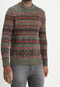 Strickpullover in Olivgrün mit roten und schwarzen geometrischen Mustern, Rundhalsausschnitt und gerippten Bündchen und Saum. Strukturierter Stoff.