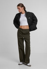 Chaqueta acolchada negra, top corto blanco, pantalones verde oliva, zapatillas beige y blancas. Corte relajado, cremallera visible y detalle de botón en los pantalones.