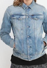 En person som bär en ljusblå denimjacka med knäppta bröstfickor och händerna i sidfickorna, svart skjorta och byxor med kamouflagemönster.