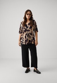 Top de manga corta y corte holgado en beige con patrón abstracto marrón oscuro; combinado con pantalones de pierna ancha negros y mocasines negros con un acento dorado.