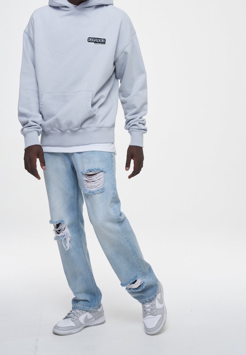 Personne portant un sweat à capuche Pegador gris clair, un jean déchiré bleu clair usé, et des baskets Nike blanc-gris, debout sur fond blanc.