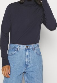 Marinefarbener Rollkragenpullover mit gerippter Textur und langen Ärmeln. Getragen mit hochgeschnittenen, hellblauen Jeans im klassischen Fünf-Taschen-Design.