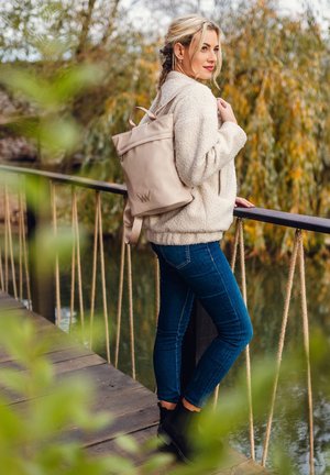 Beige backpack, minimalist design, smooth texture, logo accent; cozy white fleece jacket, dark blue jeans, black ankle boots. Natural setting.