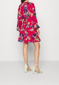 Květinové růžové šaty s dlouhými rukávy a vrstvenou sukní, s výstřihem do V a barevnými květinovými vzory. Vybavené mátově zelenými sandály na blokovém podpatku.