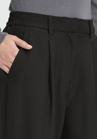 Pantalon noir sur mesure avec une taille haute, doté de plis à l'avant et de poches latérales, fabriqué dans un tissu lisse.