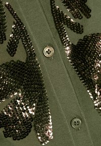 Olivgrön cardigan med paljetter i ett blommönster, fem knappstängning och en mjuk textur med synlig sömnad.