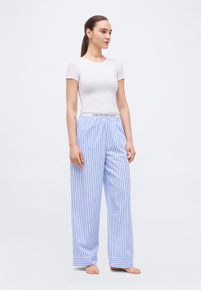 Γυναίκα με καστανά μαλλιά δεμένα πίσω, φορώντας λευκό εφαρμοστό τοπ με κοντά μανίκια και άνετο μπλε-άσπρο ριγέ παντελόνι με φαρδιές παντελόνιες, όρθια ξυπόλυτη.