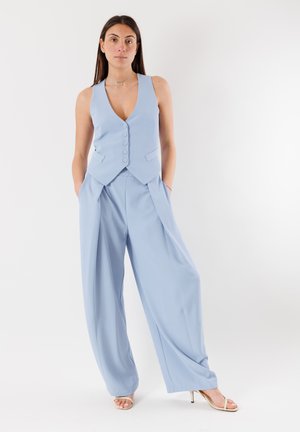 Giovane donna in piedi con le mani nelle tasche, indossa un gilet azzurro chiaro senza maniche con bottoni e pantaloni a gamba larga, tacchi beige, sfondo neutro.