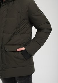 Giacca imbottita verde oliva con cappuccio, dotata di due tasche frontali, bottoni a pressione e polsini a costine. Tessuto liscio con design trapuntato.