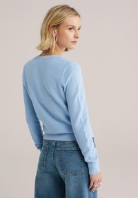 Svetlo modra pletena majica z reliefnim vzorcem, rebrastimi manšetami in robom. Kombinirana z visokim pasom modrih kavbojk iz jeansa.