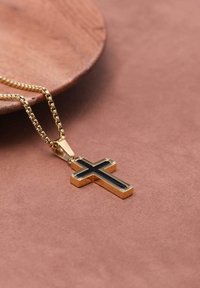 Gouden kruis hanger met een zwarte emaille inleg, bevestigd aan een delicate, glanzende ketting, rustend op een gestructureerd, zacht oppervlak.