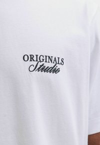 Λευκό βαμβακερό T-shirt με μαύρο κεντητό κείμενο "ORIGINALS" και "Studio." Απλό σχέδιο με κοντά μανίκια και στρογγυλή λαιμόκοψη.