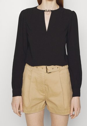 Femme portant un haut noir à manches longues avec un col en V et un short beige taille haute avec une ceinture et une boucle dorée.