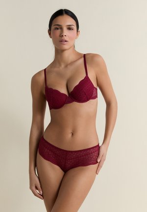 Mujer con sujetador de encaje burdeos y bragas de tiro alto a juego, de pie contra un fondo beige liso.