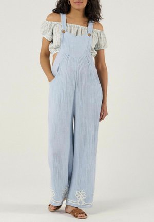 Vrouw draagt lichtblauwe wijd uitlopende tuinbroek met witte borduursels aan de zoom, gecombineerd met een off-shoulder geruit crop top en bruine sandalen.