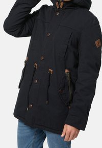 Veste noire à capuche avec fermeture éclair et boutons, poches latérales zippées, et accents en cuir marron. Tissu texturé et cordons ajustables.