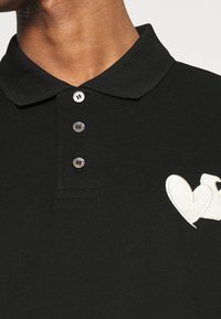 Svart polotröja med texturerad tyg, tre knappar i kragen och en vit broderad hjärtlogotyp på vänster bröst.