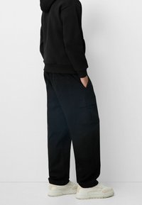 Osoba stojící s rukama v kapsách, oblečená v černých cargo kalhotách, černé mikině a bílých teniskách na světlem neutrálním pozadí.