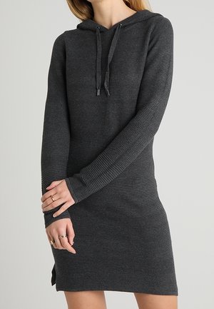 Mujer con un vestido tipo sudadera de punto gris oscuro, de manga larga y con cordones, de pie con los brazos cruzados frente a un fondo sencillo.