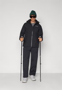 Veste de ski noire avec fermeture éclair à l'avant, associée à un pantalon noir. Les accessoires incluent un bonnet vert et des lunettes de soleil foncées. L'utilisateur tient des bâtons de trekking.