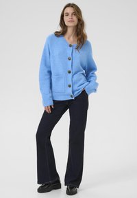 Lyseblå strikket cardigan med rund halsudskæring, fire store brune knapper, ribbede manchetter og to forlommer. Kombineret med mørke denimbukser.
