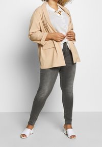 Beżowa, oversize'owa marynarka z podwiniętymi rękawami, w połączeniu z białą koszulą w kropki i szarymi, obcisłymi dżinsami. Białe klapki na odsłoniętych palcach dopełniają całość.