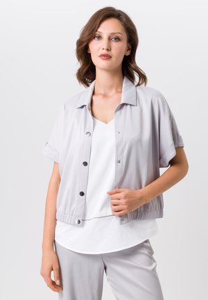 Lichtgrijze korte mouwjas met drukknoopsluitingen, elastische tailleband en een gladde textuur, gedragen over een witte top. Eenvoudig en functioneel ontwerp.