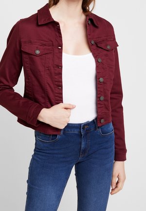 Kvinde iført en burgunderrød jakke med knapper over en hvid top og blå denimjeans, mens hun holder fast i jakkeforespidsen med den ene hånd.