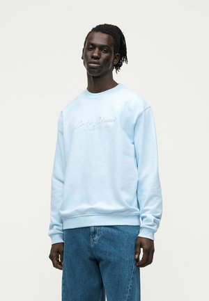 Młody mężczyzna o ciemnej karnacji i dredach, ubrany w jasnoniebieską bluzę i niebieskie dżinsy, stojący na jednolitym jasnym tle.