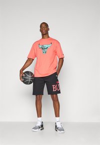 Korálová tričká s krátkym rukávom s grafikou býka; čierne kraťasy s potlačou "Chicago Bulls" v červenej farbe; šedé športové topánky; drží basketbal.
