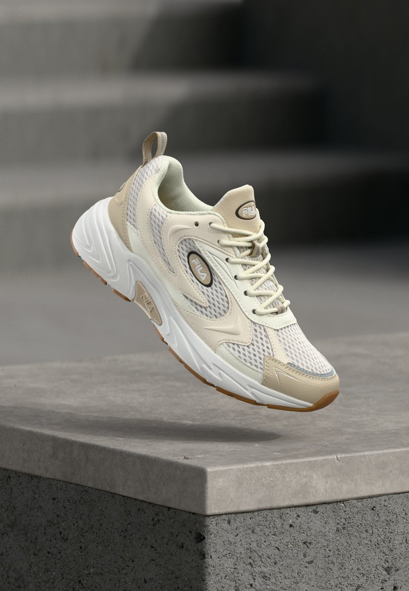 Μπεζ και λευκό αθλητικό παπούτσι FILA με λεπτομέρειες από δίχτυ και δέρμα, να αιωρείται πάνω από μια τσιμεντένια άκρη με σκάλες στο φόντο.