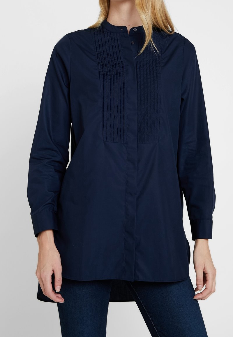 Chemise bleu marine à col rond, avec des plis verticaux sur le devant et des manches longues. Tissu lisse avec une coupe décontractée.