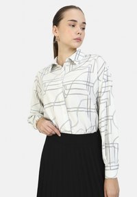 Camicia bianca con stampa geometrica nera, colletto classico e maniche lunghe, abbinata a una gonna plissettata nera.