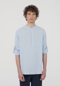 Camicia azzurro chiaro a maniche lunghe con colletto rotondo, pattina con bottoni e polsini arrotolati, realizzata in un tessuto morbido e testurizzato.