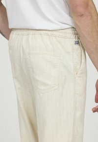 Lichtbeige gestreepte broek met een elastische tailleband, voorzien van twee zijzakken en één achterzak met een logo-label aan de achterkant.