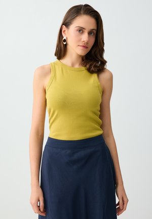 Kobieta z brązowymi włosami do ramion, nosząca złote kolczyki koła, żółtą bezrękawnikową bluzkę i granatową spódnicę, stojąca na jednolitym tle.