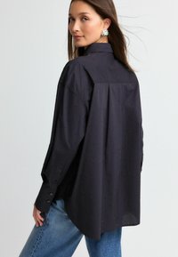 Sort skjorte med knapper og et tekstureret mønster af små nitter, lange ærmer og en buet søm, parret med lyseblå jeans.