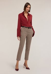 Chemise rouge à boutons avec des manches plissées, associée à un pantalon à carreaux marron et des escarpins bordeaux. Ceinture marron avec boucle en or.
