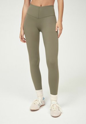 Kvinde iført olivengrønne leggings med høj talje, kombineret med hvide sokker og beige sneakers, stående på en ensfarvet hvid baggrund.