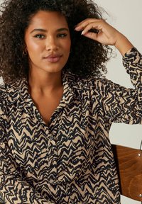 Camicia a maniche lunghe color beige con motivo a zigzag nero, dotata di colletto e chiusura frontale con bottoni. Tessuto morbido e vestibilità rilassata.