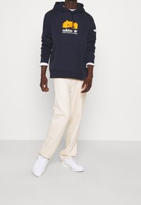 Muž v námořnickém mikině s oranžovým logem hory, bílými manžetami košile, krémových kalhotách a bílých teniskách, stojící na šedém pozadí.