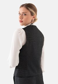 Donkere textuur vest met een getailleerde pasvorm, gedragen over een witte blouse met puffmouwen. Haar in een lage bun gestyled, met ronde oorbellen.
