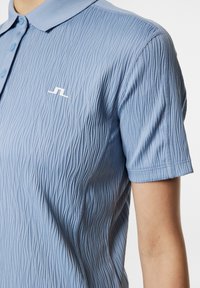 Vaaleansininen polo-paita, jossa on teksturoitu aalto-ottelu, nappilista, lyhyet hihat ja pieni valkoinen logo rinnassa.