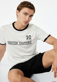 Biała koszulka z krótkim rękawem z czarnymi wykończeniami, z grafikami "10 MIAMI" i "SOCCER CULTURE". Noszona z czarnymi szortami, gładki materiał.