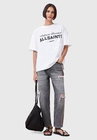 Witte oversized t-shirt met zwarte tekst, gecombineerd met versleten grijze jeans en zwarte sandalen. Accessoires zijn een zwarte totebag.