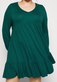 Femme portant une robe à manches longues, vert foncé, ample, avec un décolleté en V et des volants froncés à l'ourlet et au milieu.
